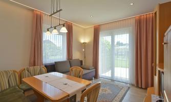 A modern living room with a dining table and cozy seating. Large windows let in plenty of daylight and provide a view of the garden.