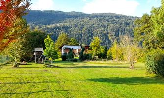 A green meadow surrounded by trees and a beautiful house. In the background, gentle mountains rise under a blue sky.