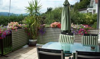 A cozy terrace with a glass table and chairs. Colorful flowers and a sun umbrella create an inviting atmosphere.