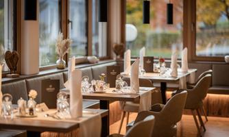 An elegant restaurant with light wooden tables and stylish chairs. It is beautifully decorated with white napkins and cozy lighting.