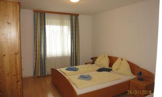 A cozy bedroom with a double bed and wooden shelves. There are large windows with curtains and fresh towels on the bed.