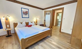 A cozy bedroom with a double bed, two nightstands, and a wooden wardrobe. The walls are light with a piece of artwork, and a carpet runs over the wooden floor.