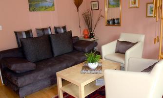 A cozy living room with a large sofa and a beige armchair. On the table, there is a plant and the room is decorated with pictures and a mirrored wall.