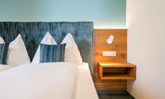 A modern bedroom with a upholstered headboard and two decorative cushions. Next to the bed is a wooden nightstand with a lamp and sockets.