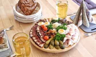 A rich cheese and sausage board with fresh vegetables, eggs, and olives. Two glasses of beer and a heart-shaped bread accompany it.