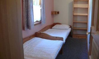 A simple bedroom with two single beds and a bookshelf. The walls are painted in soft colors and there is a window with curtains.