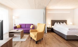 A modern room with a large bed, a purple sofa, and a yellow armchair. The room is bright and friendly with wooden flooring and plenty of natural light.