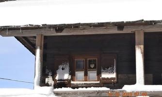 A wooden house in the snow with a window displaying a heart pendant. The surroundings are wintry and clear.
