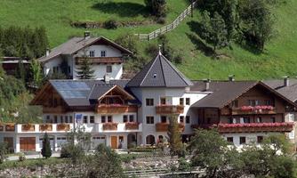 A beautiful alpine farmhouse with multiple floors and a distinctive roof. Surrounded by green hills and trees.