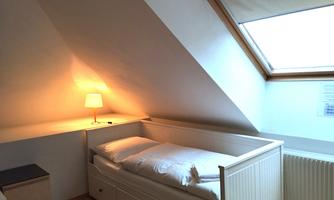 A cozy room with slanted walls and a single bed. A warm light shines from a bedside lamp next to the bed.