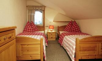 A cozy bedroom with two single beds and checked bedspreads. The room features wooden furniture and a window with a view outside.