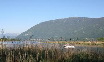 A quiet lake with reeds and a picturesque mountain landscape in the background. A small boat can be seen on the water.