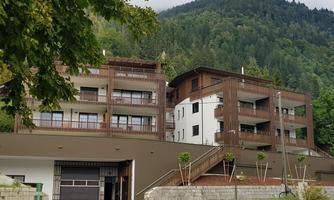 A modern building in a green landscape, surrounded by mountains. The entrance is accessible via a paved path.