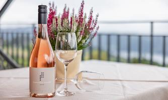 A bottle of rosé wine stands on a table next to a glass and an overturned glass. In the background, a flowering plant can be seen.
