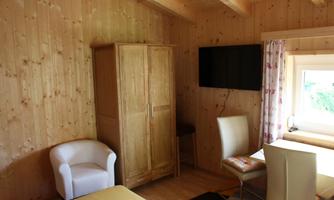 A cozy room with wooden walls and a window. There is a table, chairs, and a television on the wall.