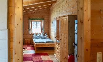 A cozy room with wooden paneling and a large window. In the background, a double bed and a wardrobe can be seen.