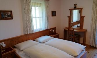 A bright bedroom with two single beds and a desk. The walls are adorned with wooden paneling and curtains, and daylight pours through the window.