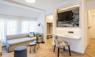A modern living room with a cozy sofa, a small table, and a television on the wall. The large windows provide plenty of daylight and a bright atmosphere.