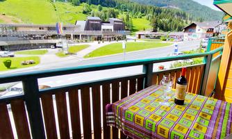 A cozy balcony with a colorful table and a glass of wine. In the background, green hills and a small village can be seen.