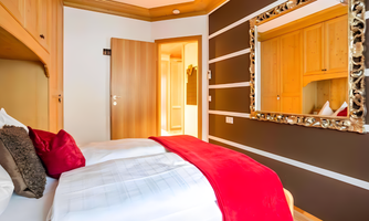A cozy bedroom with a large bed and red decorative pillows. The room has a wooden wall and an elegant mirror.
