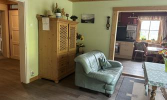 A cozy living room with a green leather sofa and a wooden cabinet. In the background, you can see a kitchen with a table and chairs.