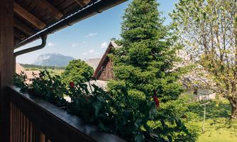 A view from the balcony of a green landscape with trees and a house in the background. The sky is clear and the mountains are visible in the distance.