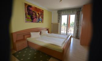 A bright bedroom with a large bed and two pillows. A colorful painting hangs on the wall, and large windows provide a view outside.