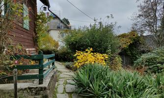 A beautiful garden with colorful plants and a paved path. In the background, there is a wooden house under a gray sky.