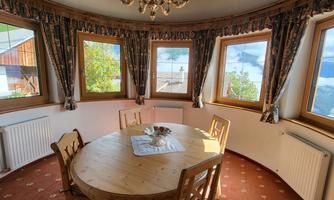 A cozy dining area with a round wooden table and four chairs. Surrounding it are large windows that offer a view of the surroundings.