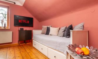 A cozy room with a pink wall and a bed with pillows. A television with the Netflix logo hangs on the wall.
