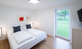 A modern bedroom with a double bed and decorative pillows. Large windows let in plenty of daylight and offer a view of the green surroundings.