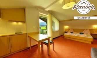 A bright guest room with a table and chairs. In the background, a bed and large windows with a view of the landscape can be seen.