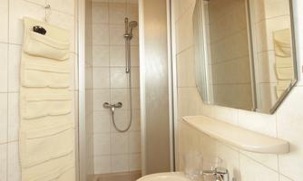 A simple bathroom with a shower, a sink, and a mirror. The walls are tiled and the wooden ceiling creates a warm atmosphere.
