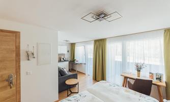 A bright room with two beds, a cozy sofa, and a small table. Large windows provide plenty of daylight, and the green curtains create an inviting atmosphere.