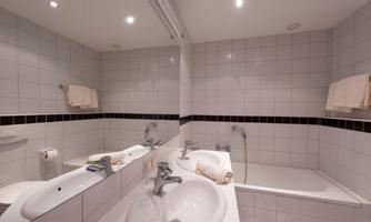 A modern bathroom with white tiles and a bathtub. There are two sinks and bright lighting.