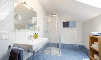 A modern bathroom with a shower and a sink. The walls are tiled white, and the floor has a blue tone.