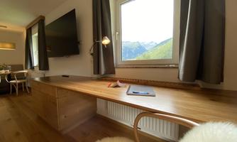 A modern room with a wooden desk and a large window through which you can see the mountains. A television hangs on the wall, and the curtains are elegant and simple.