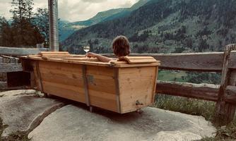 A wooden bathtub on a rock overlooking a green mountain landscape. A person is sitting relaxed in it holding a glass.