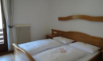 A cozy bedroom with a large wooden bed and two pillows. The room has a simple, bright decor and a window with curtains.