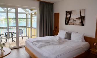 A bright, modern bedroom with a large double bed and a view of a lake. The room has a terrace with chairs and a pleasant, inviting style.