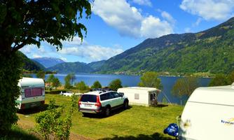 A picturesque campsite by the lake with caravans and green meadows. In the background, majestic mountains and a blue sky can be seen.