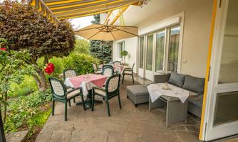 An inviting sunny terrace with a table, chairs, and a seating area under a parasol. Surrounded by green plants and a cozy atmosphere.