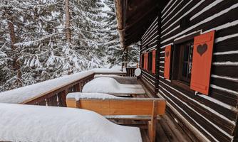 A cozy wooden cabin in the snow with orange window shutters. The balcony is covered with a thick layer of snow and surrounded by snow-covered trees.