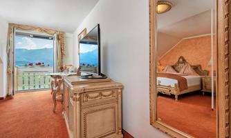 An elegant room with a television, a vanity table, and a large mirror. Through the window, you can see the mountains and a balcony with flowers.