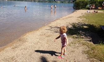 A small boy stands at the shore of a calm body of water. In the background, other people are in the water and a beautiful landscape with mountains can be seen.