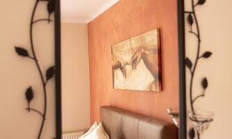 A decorative mirror with a black frame and leaf motifs. In the background, there is a relaxing bedroom featuring a picture and a cozy bed.