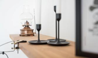 An elegant wooden shelf with a traditional oil lamp and two black candle holders. The wall in the background is simple and white.