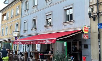 A cozy café with a terrace is located on a street. The facade of the building is painted in soft colors.
