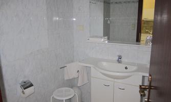 A simple bathroom with white tiles and a sink. The room contains a stool and towels are neatly hung up.
