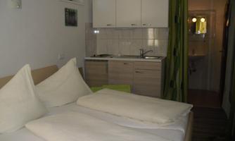 A simple bedroom with a double bed and a small kitchenette. The room has bright walls and a cozy, inviting character.